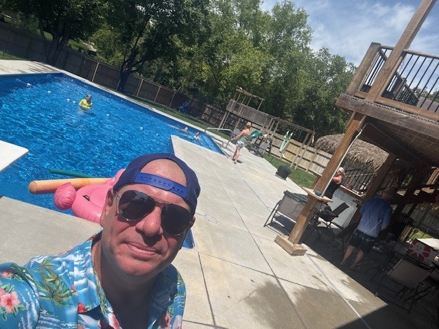 Andrew Sherwood in sunglasses and a summer shirt takes a selfie by his backyard pool where his family is enjoying the day