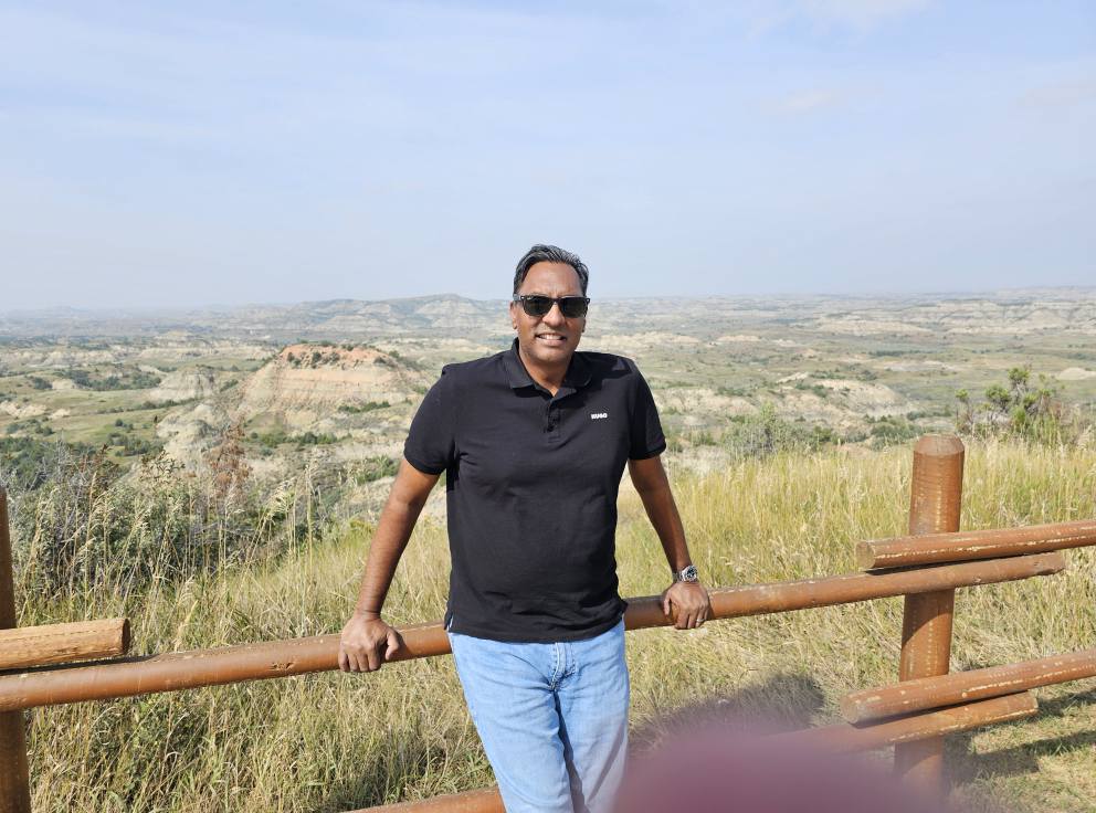Ravi Shivdat leaning against a wooden fence with the vast North Dakota Badlands behind him on a beautiful summer day