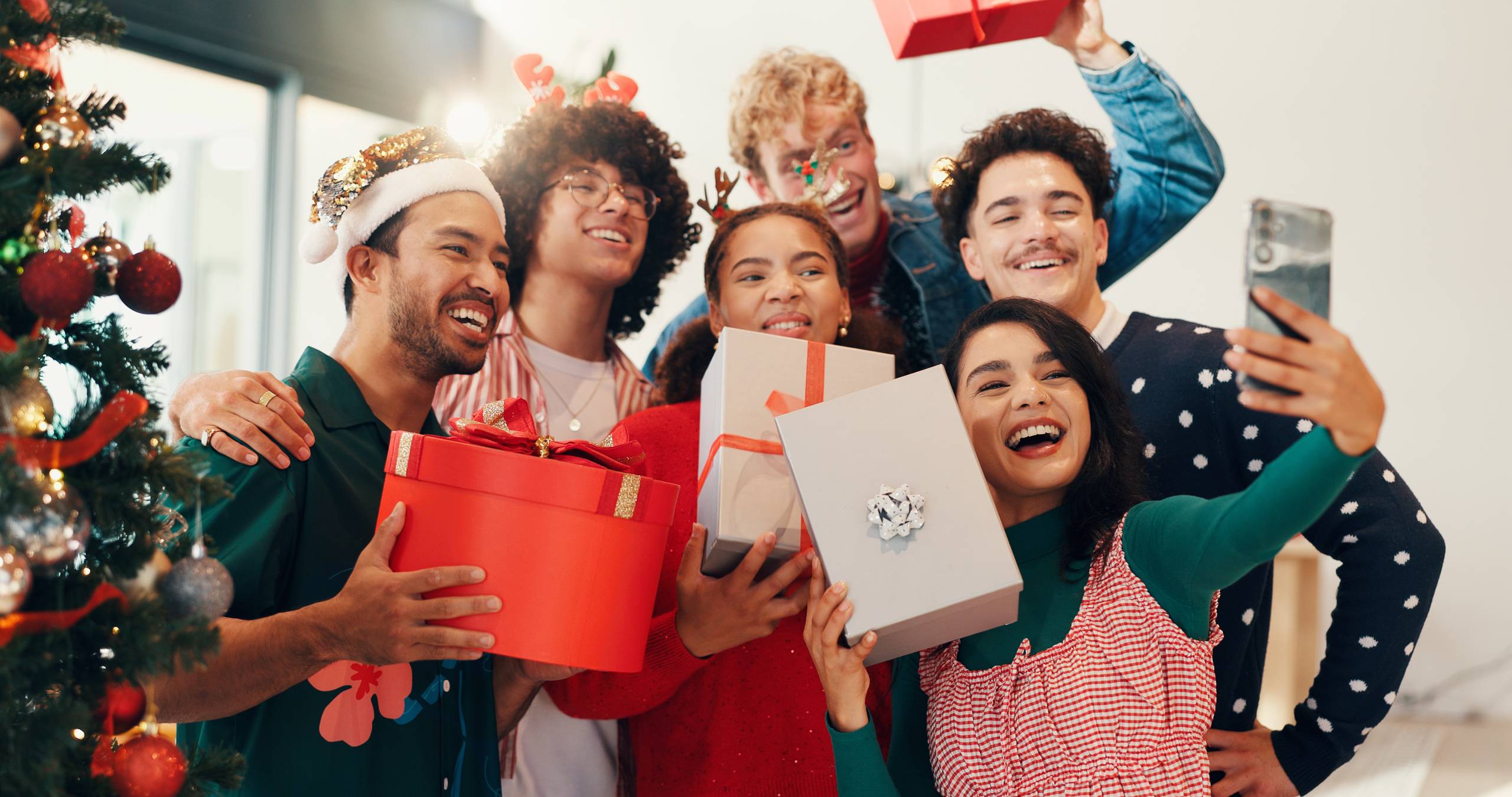 a group of festive and happy friends taking a selfie while exchanging holiday presents near a Christmas tree