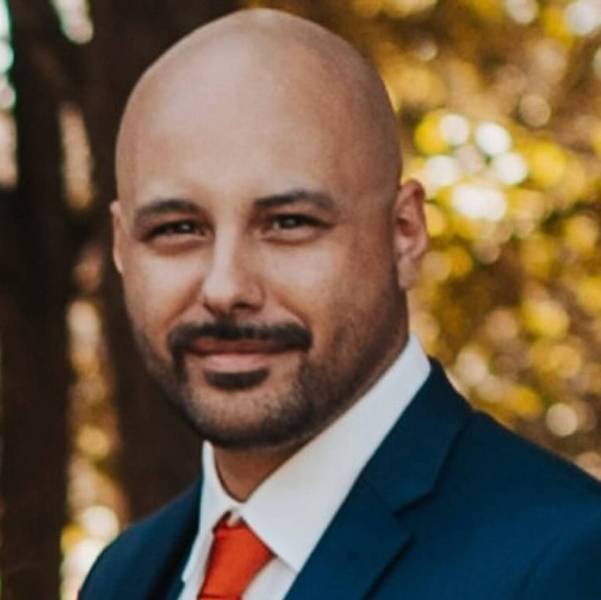 A professional headshot of Jared Lemieux standing in front of a tree while wearing a blue suit, white shirt, and orange tie